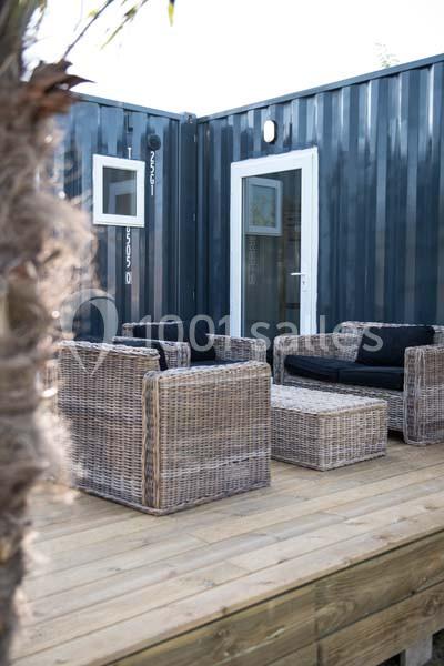 Terrasse en bois avec fauteuils en rotin et table basse, devant un bâtiment en conteneur métallique bleu.