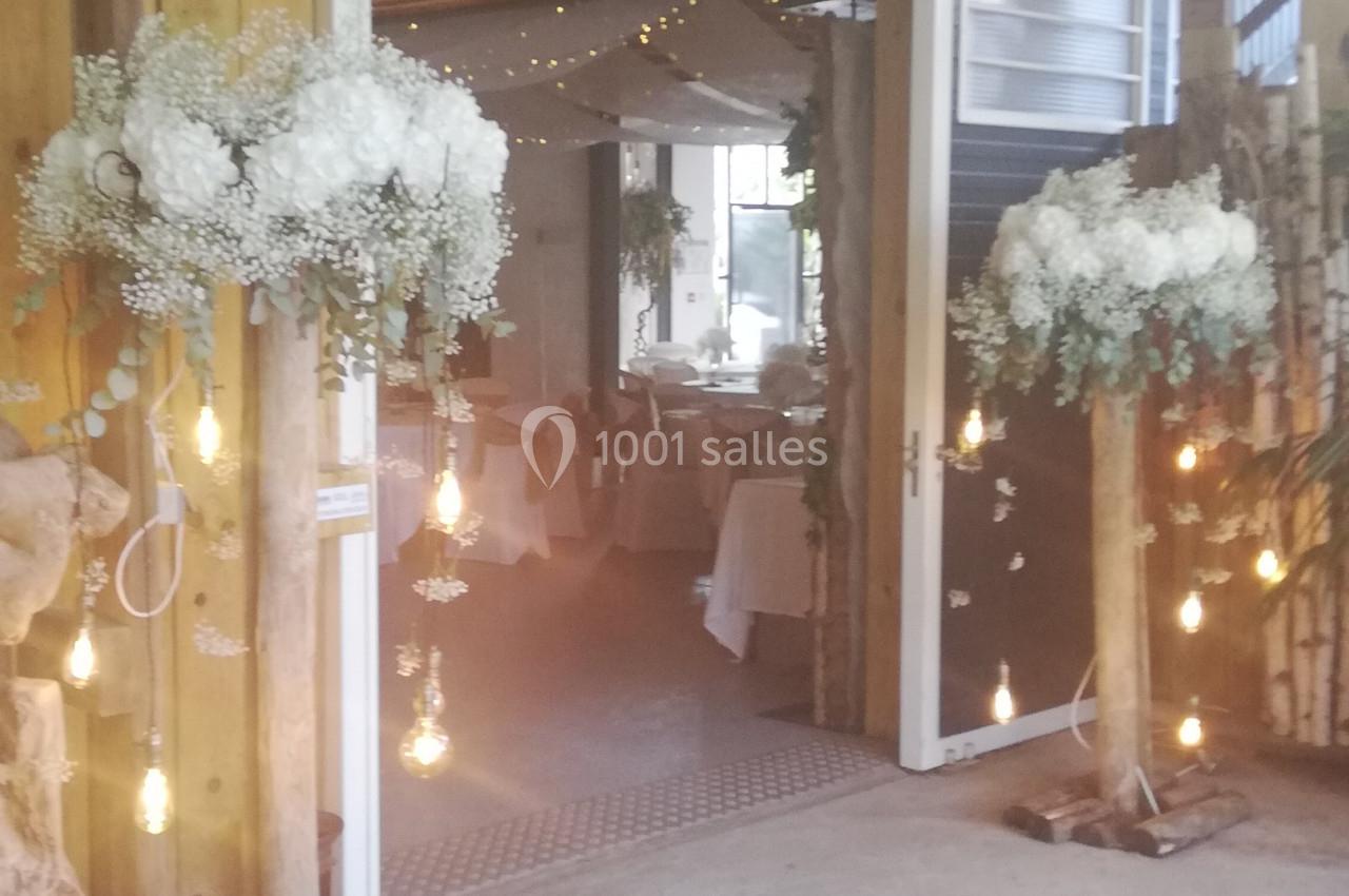 Entrée décorée avec des guirlandes lumineuses, des fleurs blanches et des tables dressées visibles à l'intérieur.