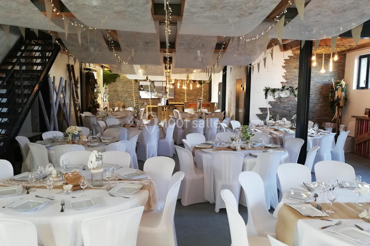 Salle de réception décorée avec des tables rondes nappées de blanc, guirlandes lumineuses et ambiance rustique.