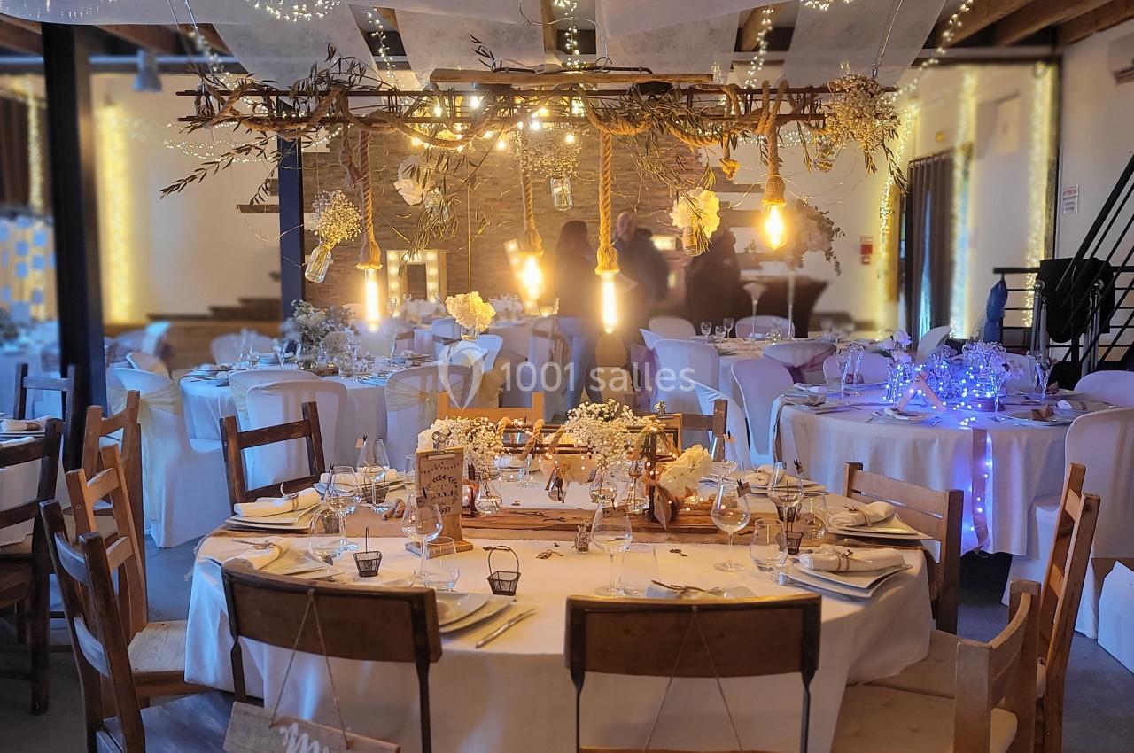 Salle de réception décorée pour un mariage avec guirlandes lumineuses, tables rondes dressées et chaises en bois.
