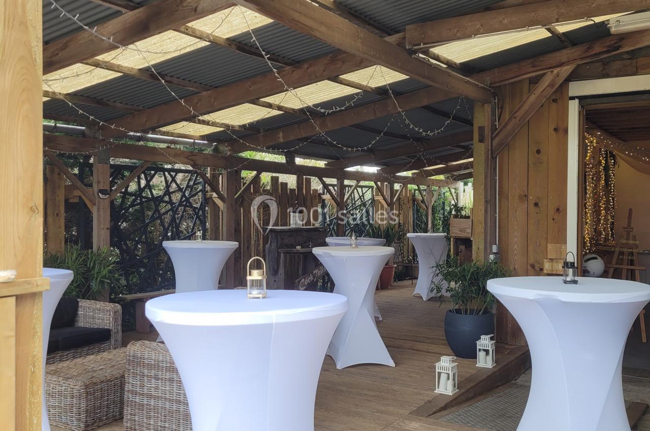 Salle en bois avec tables hautes recouvertes de nappes blanches, décorée de plantes et guirlandes lumineuses.
