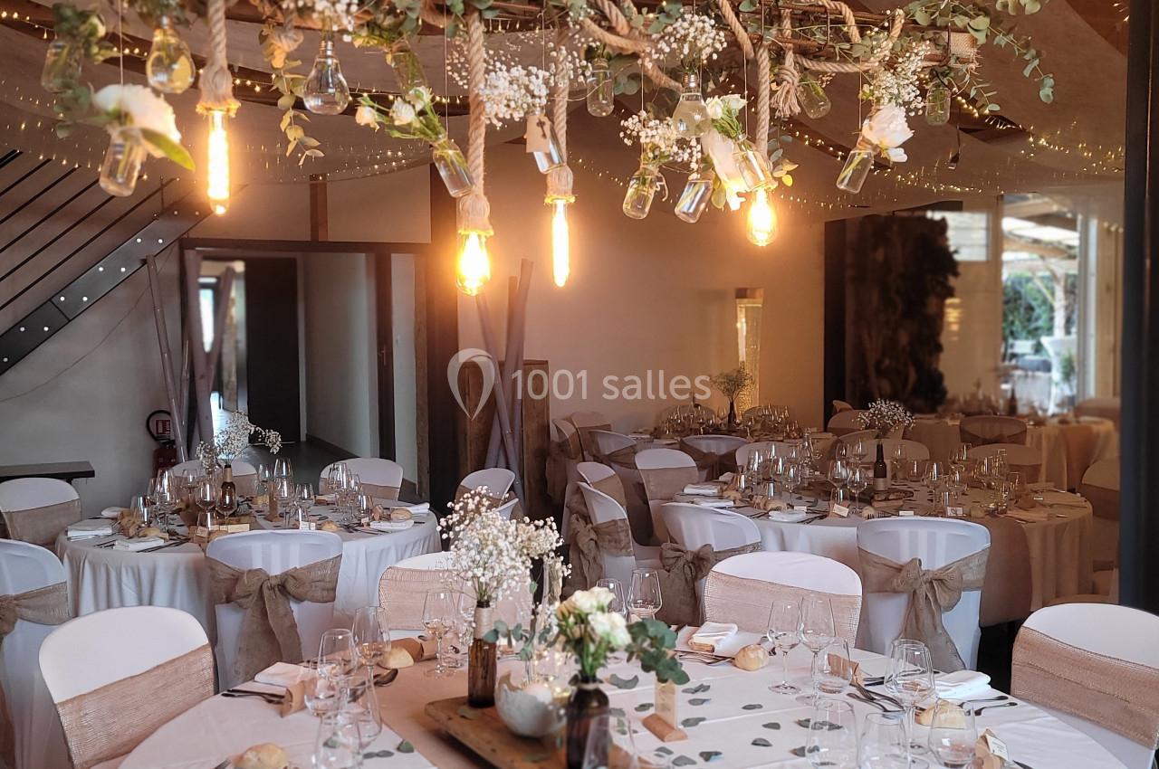Salle de réception décorée avec des tables rondes, nappes blanches, centres de table fleuris et guirlande lumineuse…