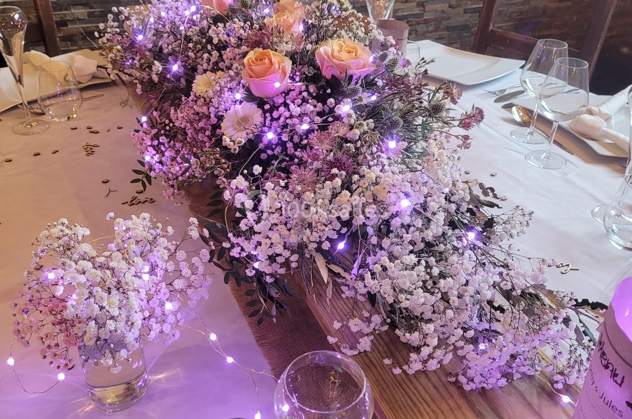 Centre de table floral avec gypsophiles, roses et guirlandes lumineuses violettes sur une table dressée.