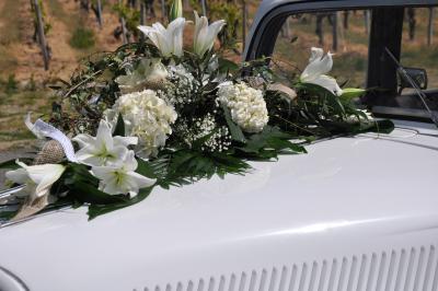 Voiture ancienne blanche décorée de fleurs blanches, stationnée sur un chemin près de vignes et d'un arbre.