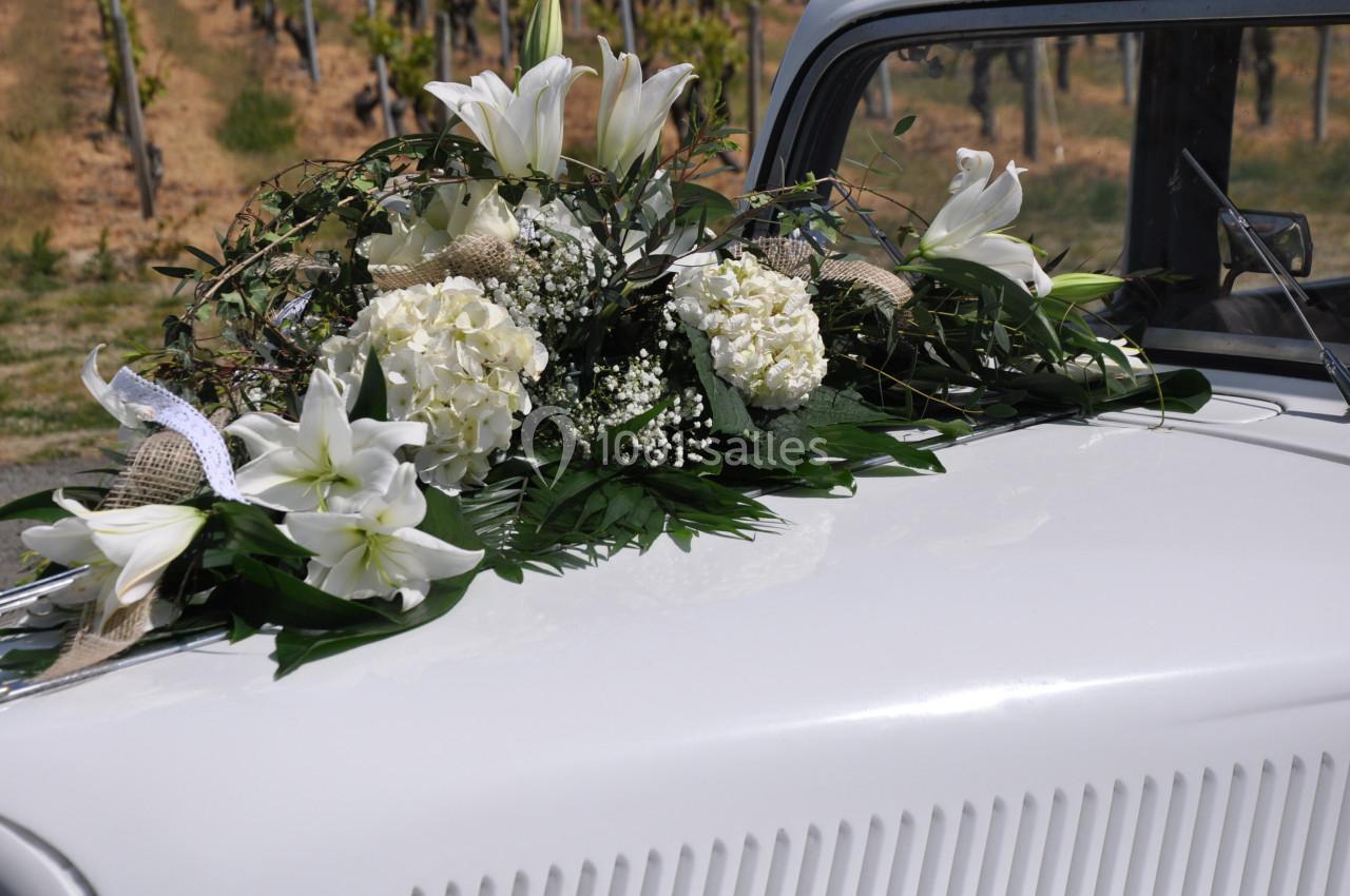 Arrangement floral blanc avec des lys et des hortensias posé sur le capot d'une voiture ancienne dans un vignoble.