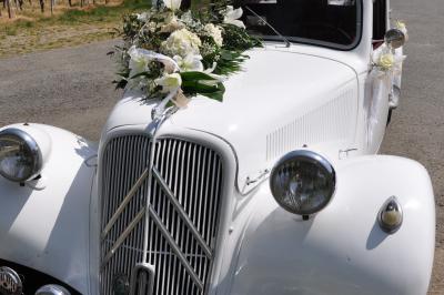 Voiture ancienne blanche décorée de fleurs blanches, stationnée sur un chemin près de vignes et d'un arbre.