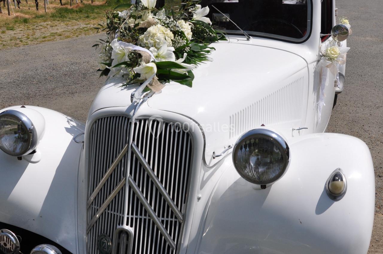 Voiture ancienne blanche décorée de fleurs blanches, stationnée sur un chemin près de vignes et d'un arbre.