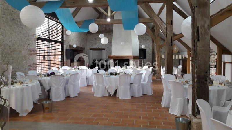 Salle de réception décorée avec des tables rondes, nappes blanches, lanternes suspendues et poutres apparentes.