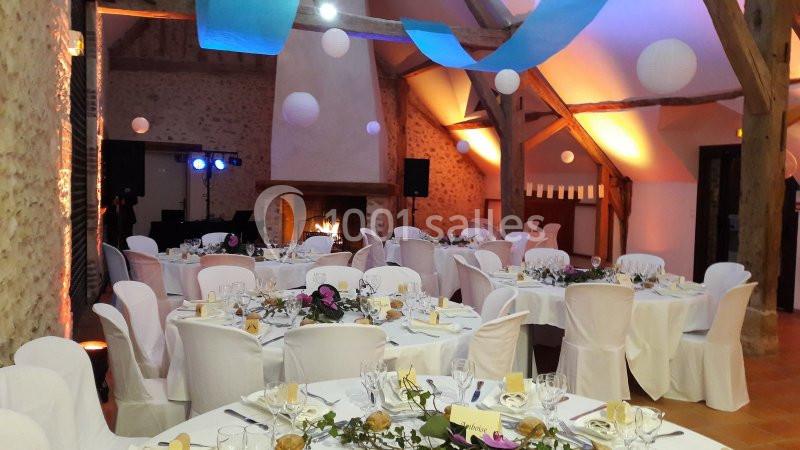 Salle de réception décorée avec des tables rondes, nappes blanches, guirlandes lumineuses et cheminées en pierre.
