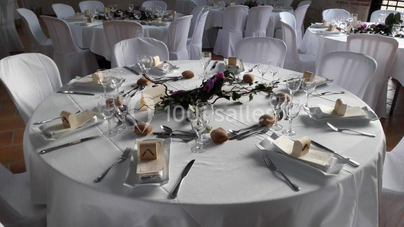 Table ronde dressée pour un repas, avec nappes blanches, couverts, verres, menus et décoration florale au centre.