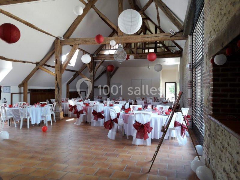 Salle de réception décorée pour un événement, avec tables rondes, nappes blanches, nœuds rouges et lanternes suspendues.