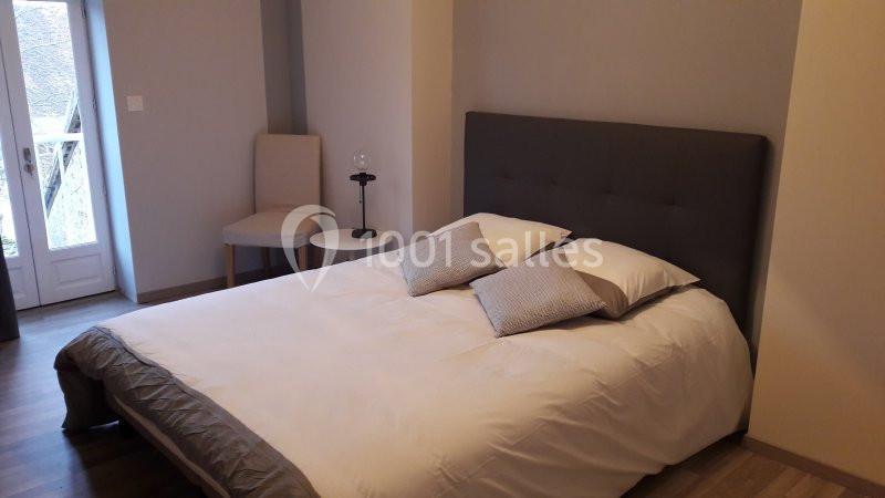 Lit double avec tête de lit grise, draps blancs et coussins, dans une chambre lumineuse avec chaise et petite table.