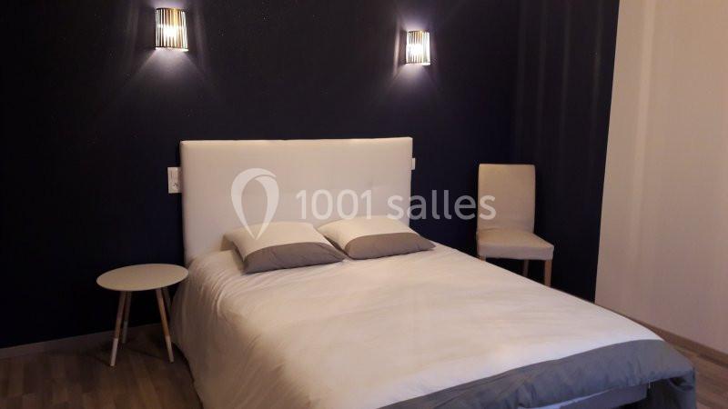 Lit double avec tête de lit blanche, deux coussins, table de chevet et chaise dans une chambre aux murs sombres.