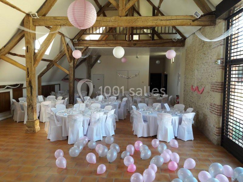 Salle de réception décorée avec des ballons roses et blancs, des tables dressées et des poutres apparentes en bois.
