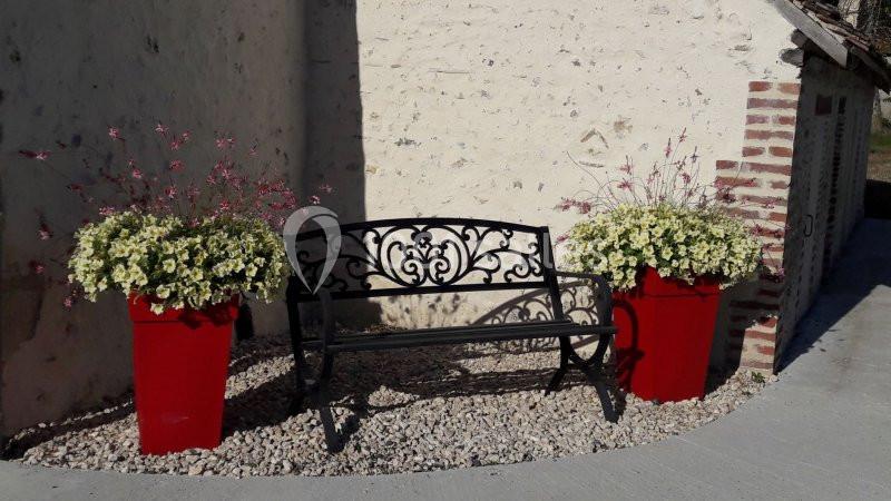 Banc en fer forgé noir entouré de deux pots rouges avec des fleurs, placé devant un mur en pierre claire.