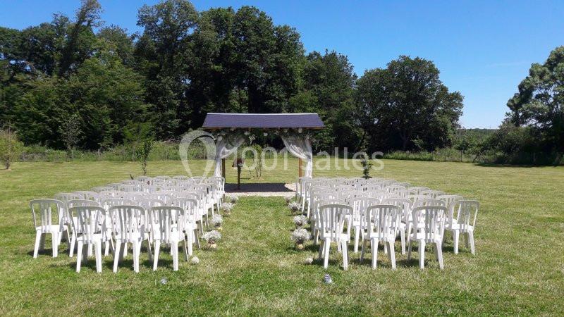 Chaises blanches disposées en rangées devant une arche décorée, installées sur une pelouse entourée d'arbres.