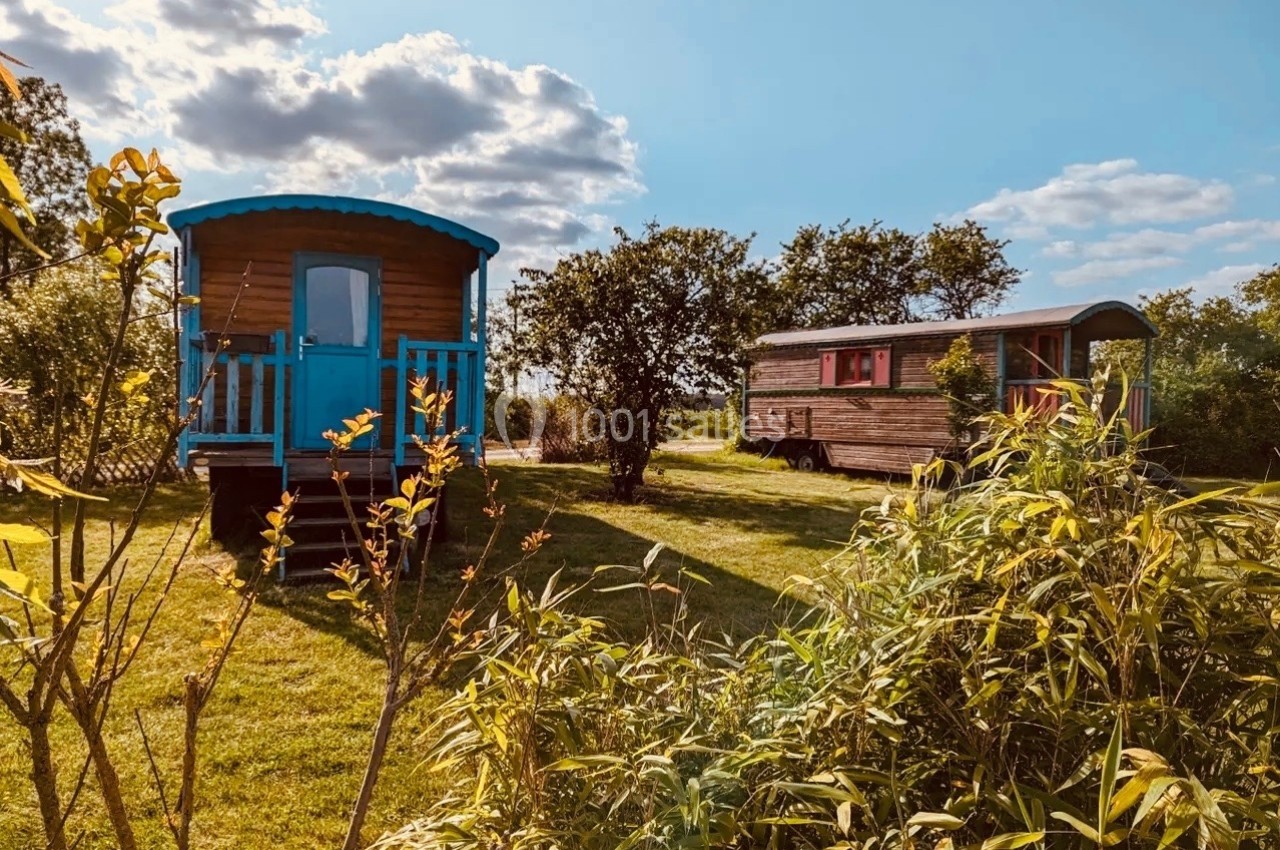 Deux roulottes en bois dans un cadre verdoyant, entourées d'arbustes et d'arbres sous un ciel partiellement nuageux.