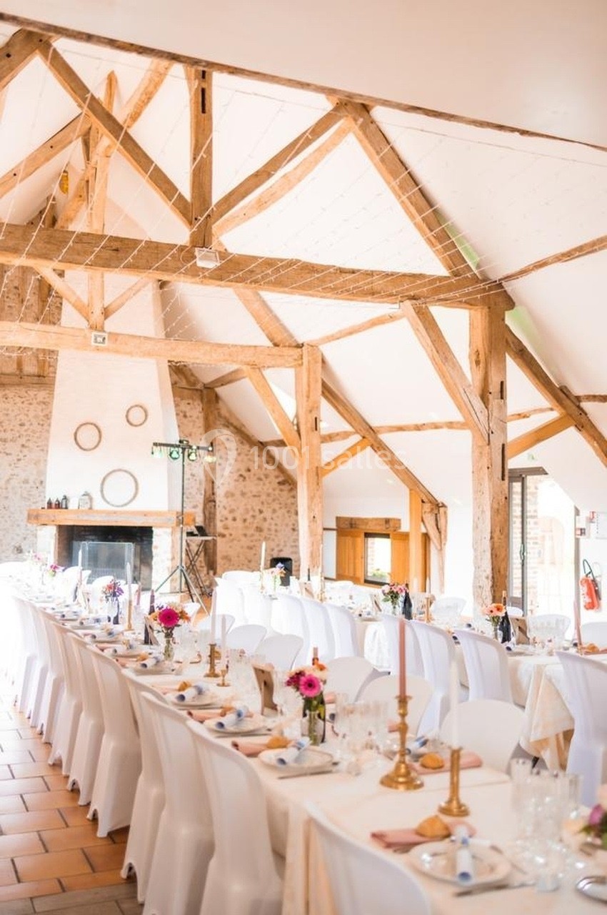 Salle de réception lumineuse avec poutres apparentes, tables décorées de nappes blanches et bougeoirs dorés.