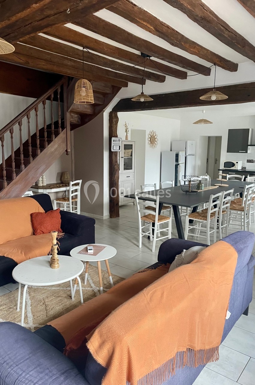 Salon spacieux avec poutres apparentes, canapés orange, table à manger et cuisine ouverte en arrière-plan.