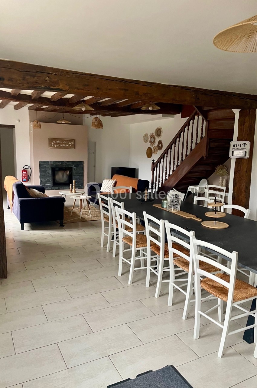 Salle à manger avec une grande table et des chaises blanches, ouverte sur un salon avec cheminée et escalier en bois.