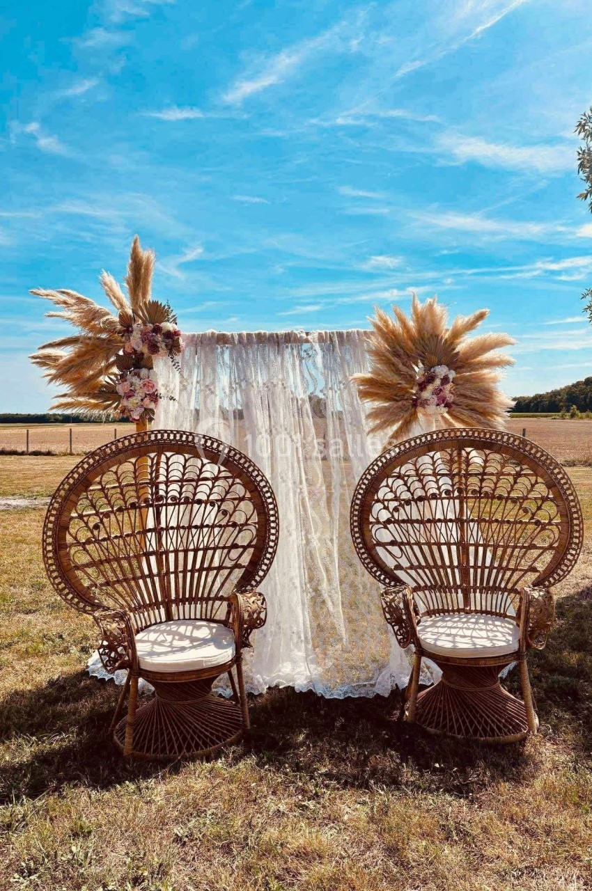 Deux fauteuils en rotin devant un rideau en dentelle décoré de fleurs et de pampas, installés dans un champ ensoleillé.