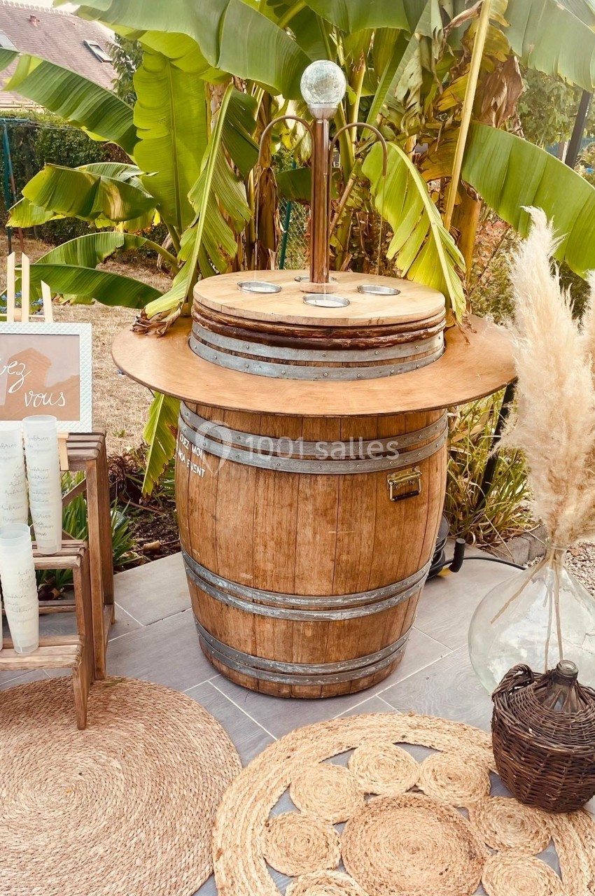 Tonneau en bois transformé en bar avec décoration végétale, verres et pancarte ’Servez-vous’ dans un jardin.