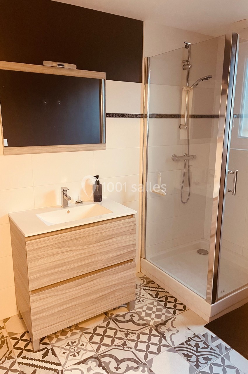 Salle de bain moderne avec meuble en bois clair, miroir, douche vitrée et carrelage à motifs géométriques.