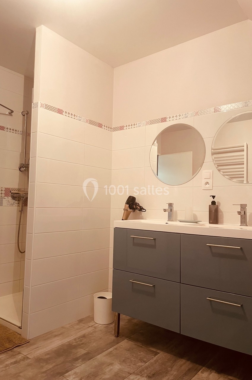 Salle de bain avec douche, meuble à double vasque bleu, miroirs ronds et carrelage mural blanc.