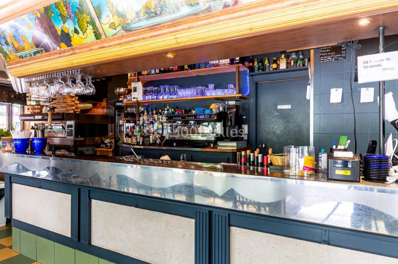 Comptoir de bar avec étagères garnies de verres, bouteilles et machine à café, dans un décor coloré et lumineux.