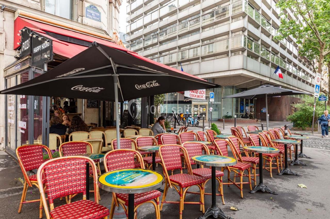 Terrasse de café parisien avec des chaises en osier rouge et des tables rondes, située au bord d'une rue urbaine.