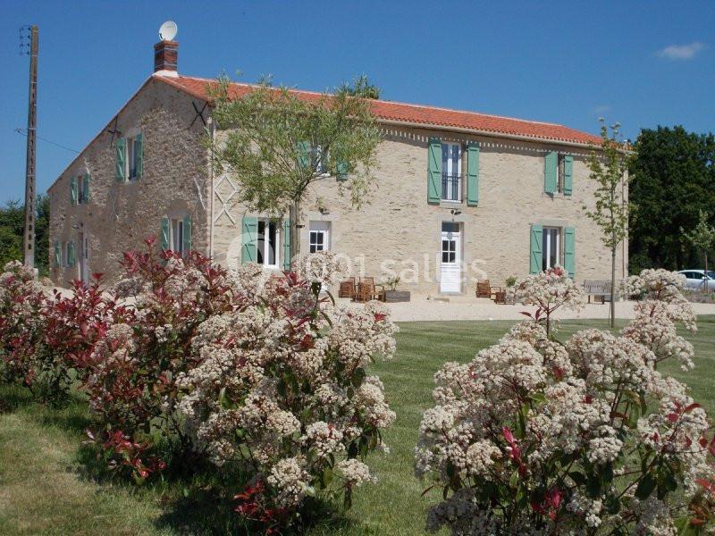 Maison en pierre avec volets verts, entourée d'un jardin fleuri et d'arbustes, sous un ciel bleu dégagé.