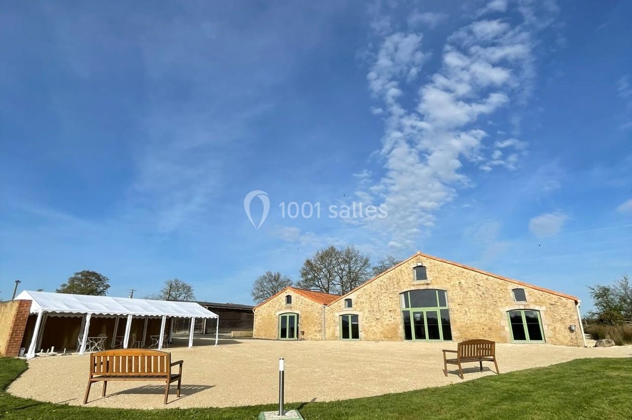 Bâtiments en pierre avec grandes fenêtres, terrasse aménagée et pelouse sous un ciel bleu partiellement nuageux.