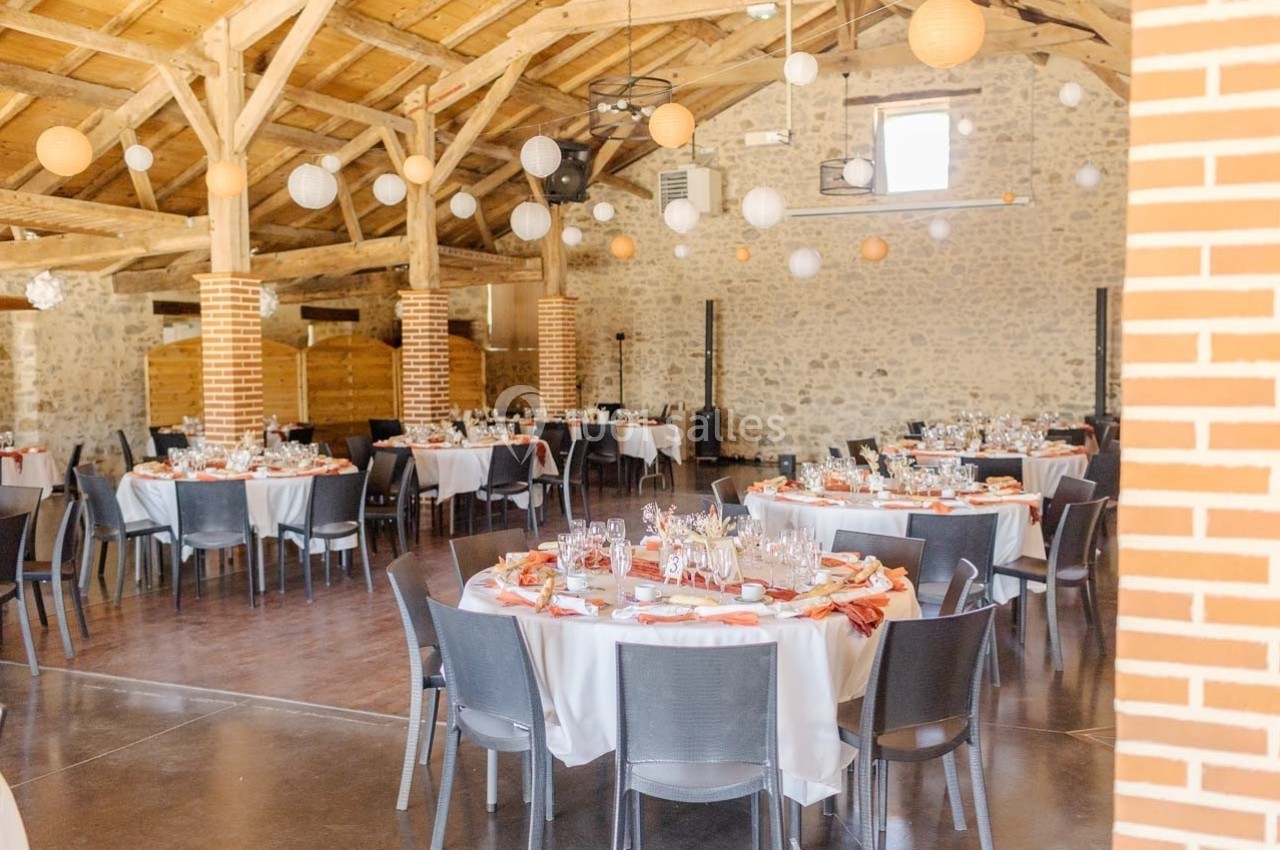Salle de réception avec des tables rondes dressées, décorée de lanternes suspendues et de murs en pierre apparente.