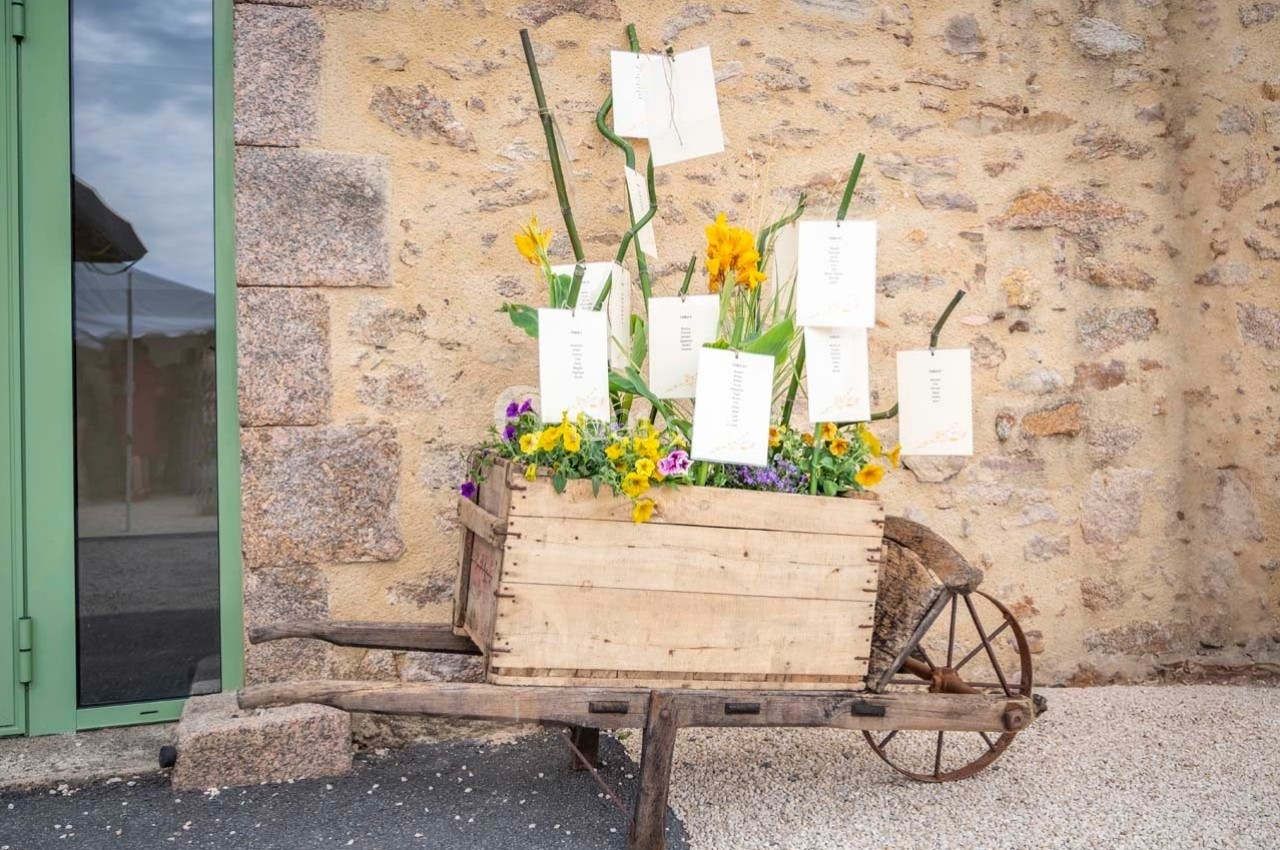 Charrette en bois remplie de fleurs colorées et de cartes suspendues, placée devant un mur en pierre.