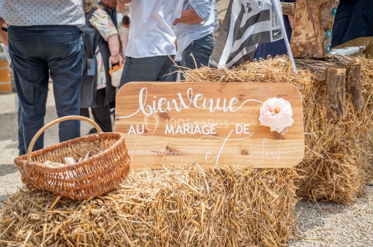 Panneau en bois ’Bienvenue au mariage de’ posé sur des bottes de paille, décoré d'une fleur et d'un panier en osier.