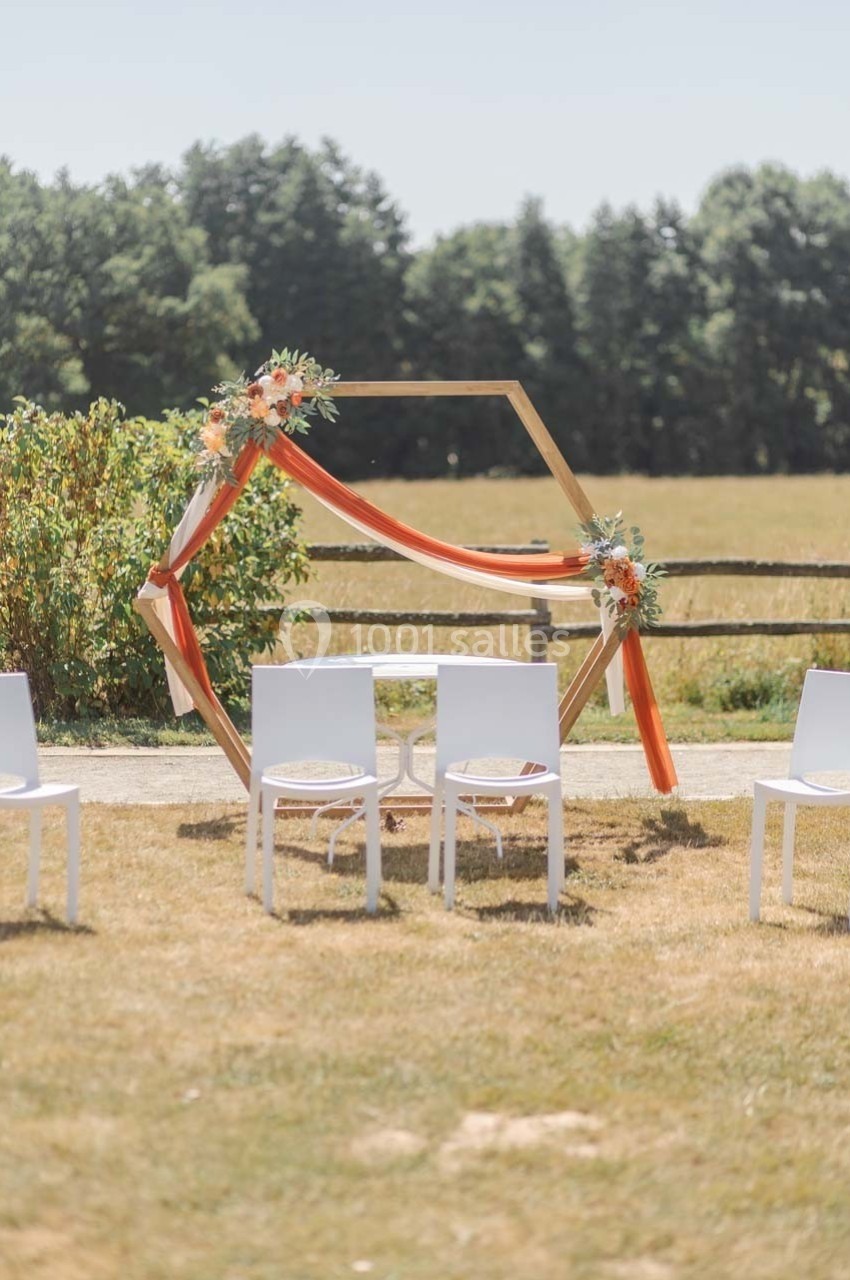 Arche hexagonale décorée de fleurs et de tissus orange, entourée de chaises blanches dans un cadre champêtre.