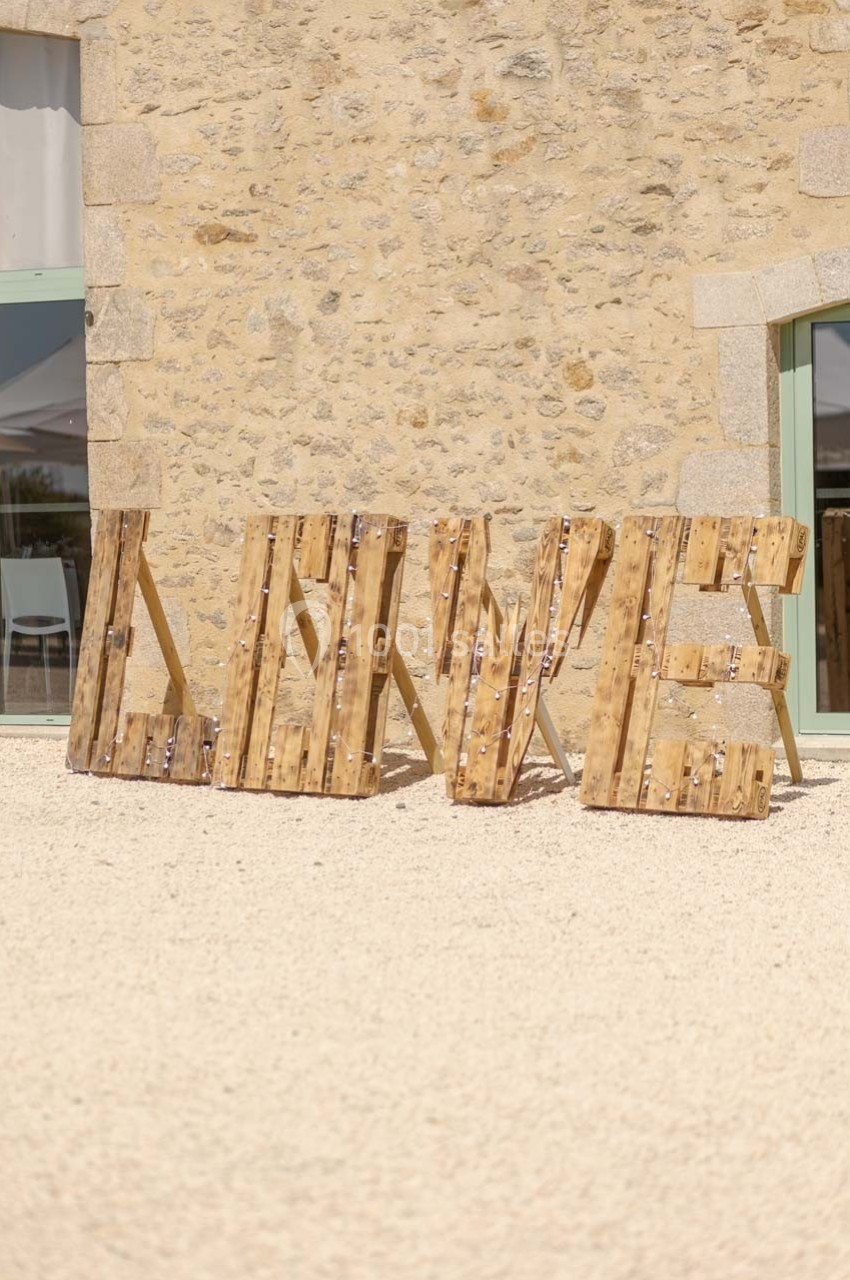 Lettres en bois formant le mot ’LOVE’ disposées devant un mur en pierre, sur un sol clair.