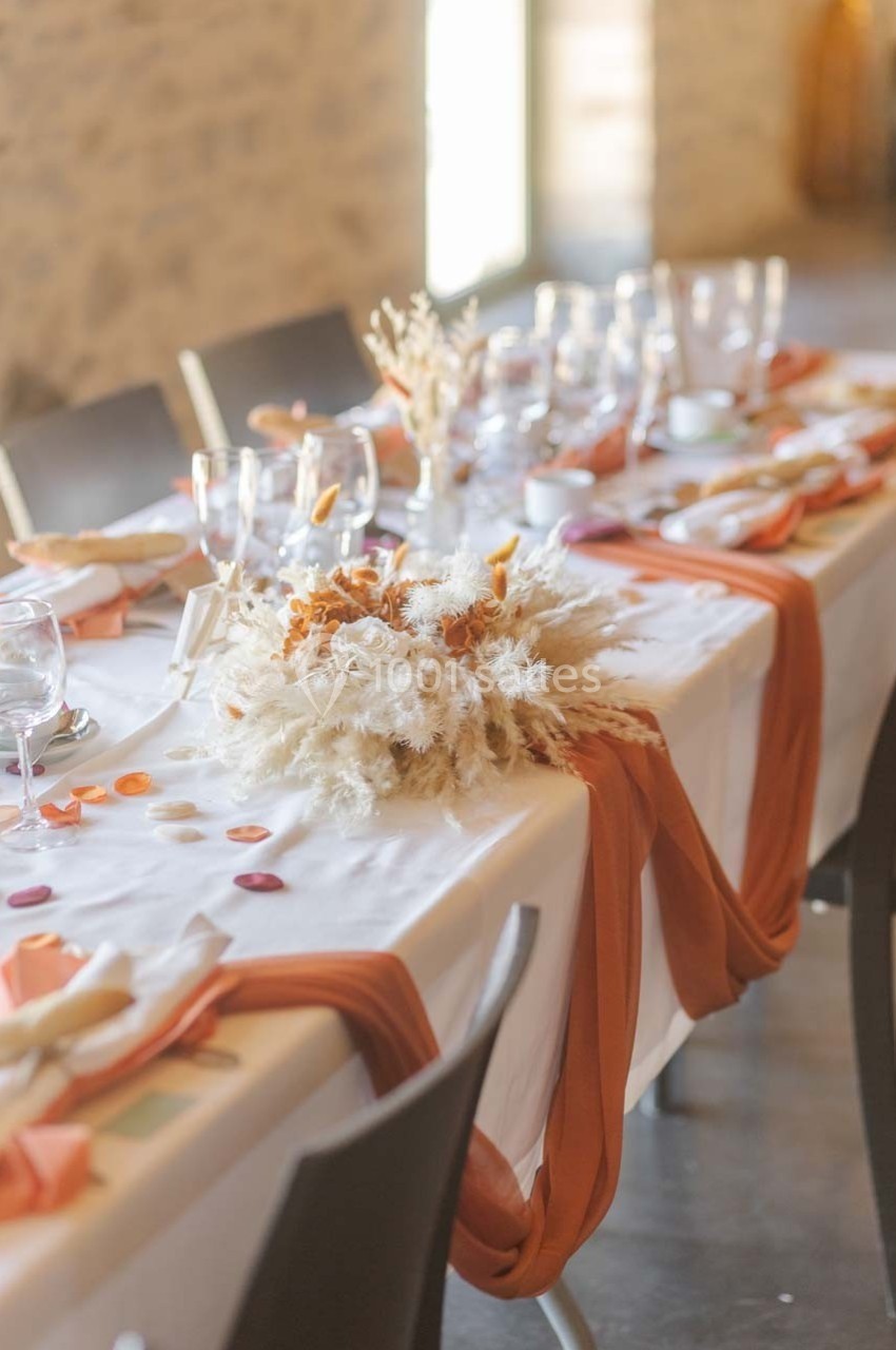 Table décorée avec une nappe blanche, des chemins de table orange, des verres et un centre de table en fleurs séchées.