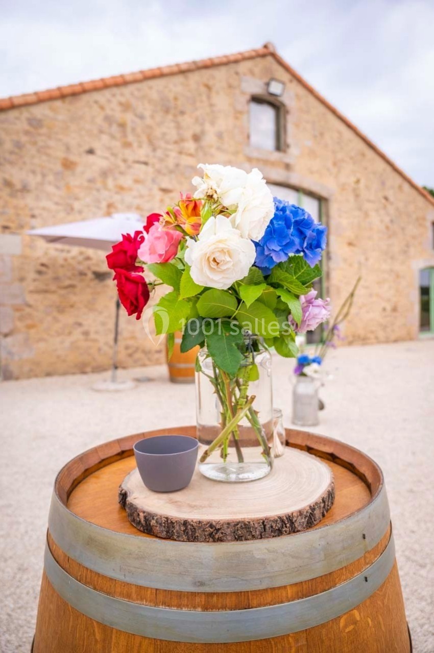 Bouquet de fleurs colorées dans un vase en verre posé sur un tonneau en bois devant un bâtiment en pierre.