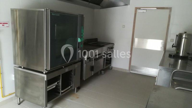 Cuisine professionnelle avec four, plaques de cuisson, plan de travail en inox et porte au fond de la pièce.