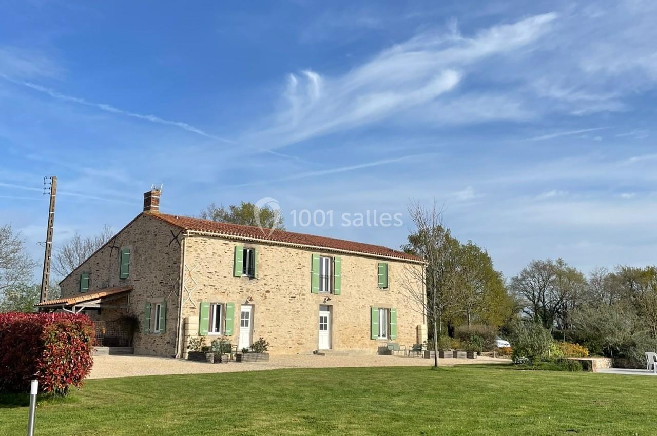 Maison en pierre avec volets verts, entourée d'un grand jardin et d'arbres, sous un ciel bleu dégagé.