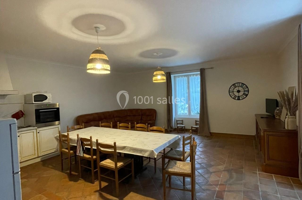 Salle à manger avec une grande table entourée de chaises, carrelage au sol, cuisine équipée et salon en arrière-plan.