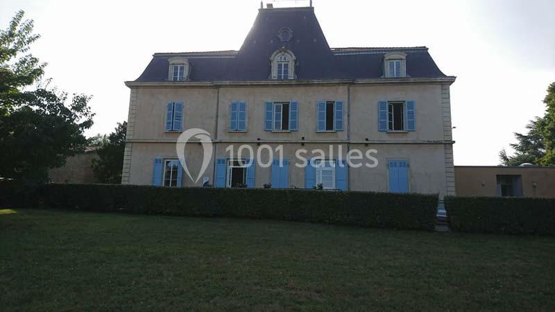 Façade d'un grand bâtiment ancien avec volets bleus, entouré d'une haie et d'une pelouse.