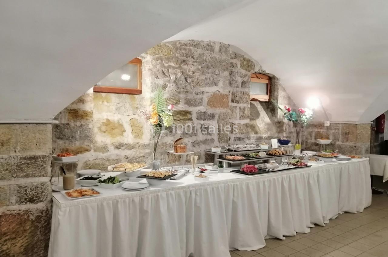 Buffet varié disposé sur une longue table drapée de blanc, dans une salle voûtée en pierre.