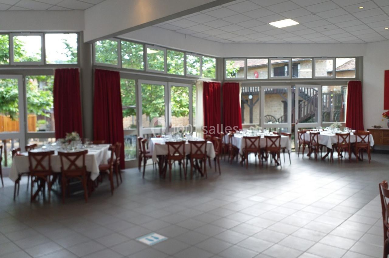 Salle lumineuse avec de grandes baies vitrées, tables rondes dressées avec nappes blanches et rideaux rouges.