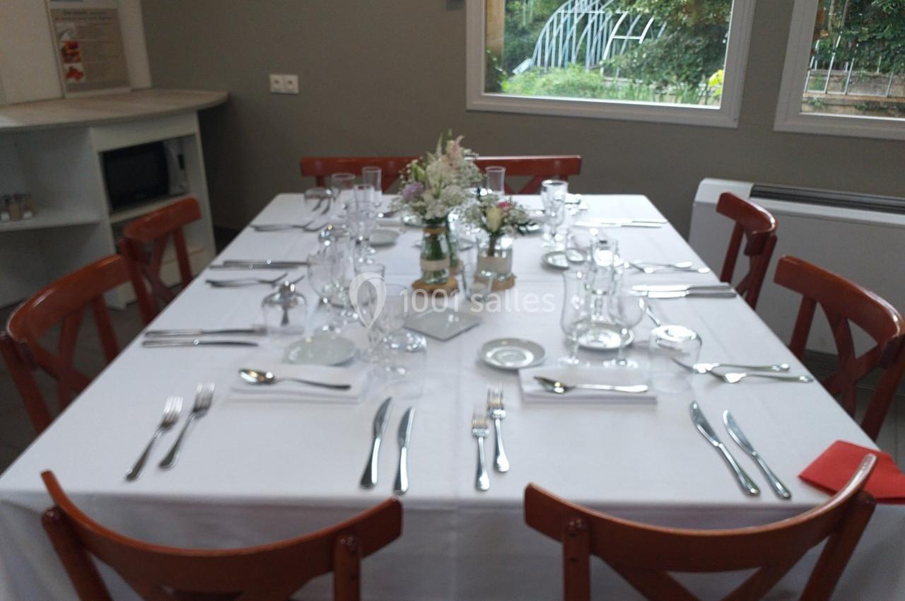 Table dressée avec nappe blanche, couverts, verres et décoration florale, entourée de chaises en bois dans une salle…