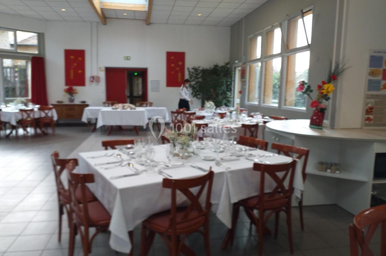 Salle de restaurant lumineuse avec tables dressées de nappes blanches, chaises en bois et décorations florales.