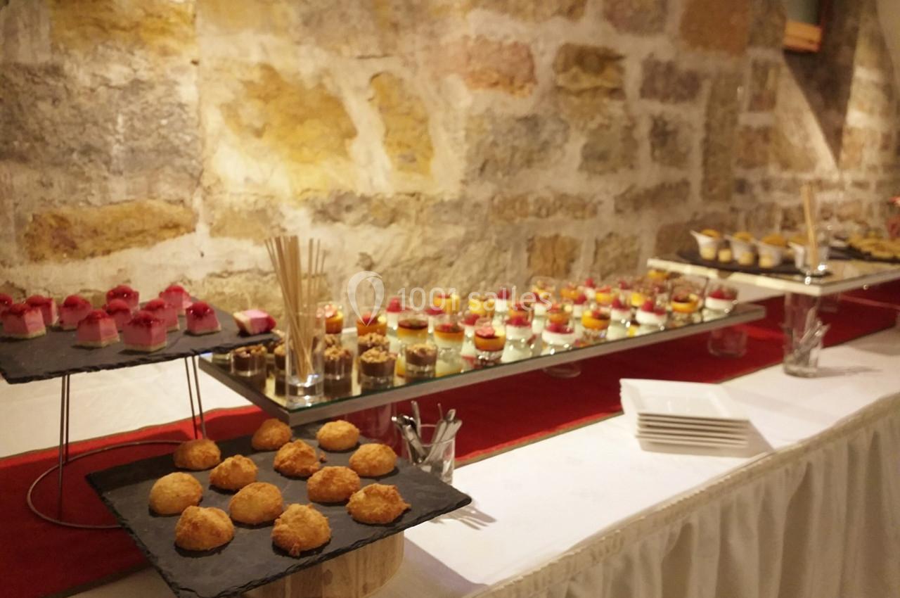 Buffet de desserts variés disposés sur des plateaux devant un mur en pierre, avec nappes blanches et décoration sobre.