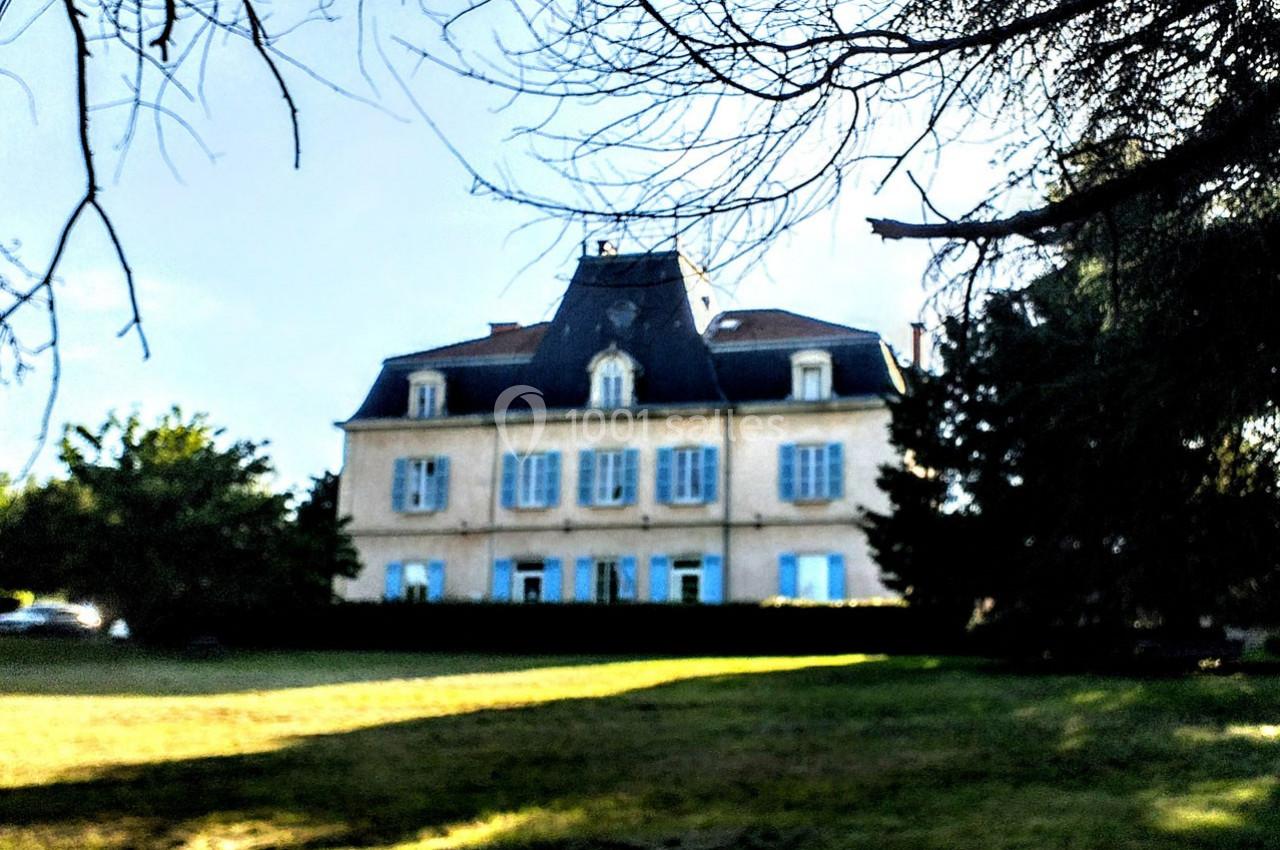 Grande maison ancienne à trois étages avec des volets bleus, entourée d'arbres et d'une pelouse ensoleillée.