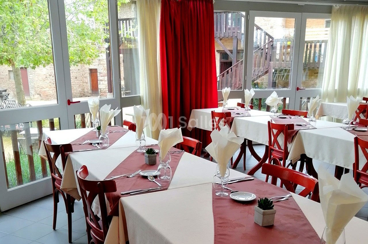 Salle de restaurant lumineuse avec tables dressées, nappes blanches et rouges, et vue sur une cour extérieure.